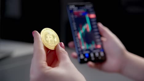 Eine-Frau-Untersucht-Die-Physische-Goldene-Bitcoin-Münze,-Während-Sie-Die-Kryptowährungs-Handels-App-Auf-Ihrem-Smartphone-überprüft