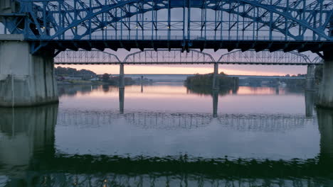 Aerial-footage-during-sunrise-close-to-the-water-of-the-Tennessee-River-going-under-the-Market-Street-Bridge-during-sunrise-with-yellow-and-purple-in-the-clouds