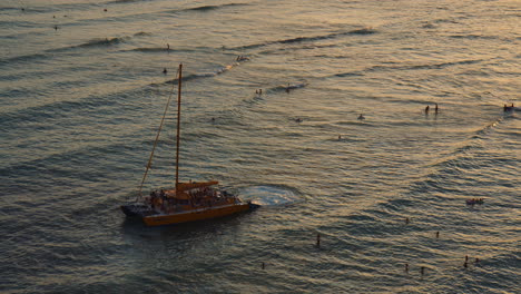 Yellow-Catamaran-Setting-Sail-at-Golden-Sunset-in-Shallow-Water-With-Tourists-Enjoying-Waves,-Hawaii