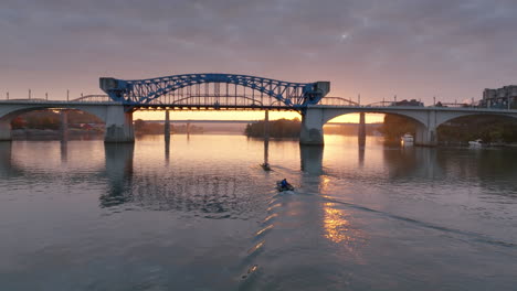 Aerial-footage-during-sunrise-close-to-the-water-of-the-Tennessee-River-following-rowers-on-a-boat-going-under-the-Market-Street-Bridge-during-sunrise