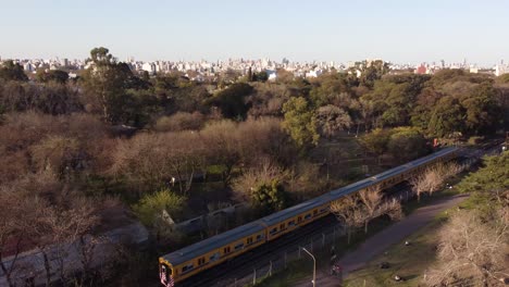 Eine-Dynamische-Aufsteigende-Luftaufnahme-Eines-Sich-Bewegenden-Gelben-Zugs,-Der-Durch-Gemüsefelder-Und-Den-öffentlichen-Agronomia-park-In-Buenos-Aires-Fährt