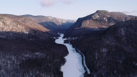 Epic-backwards-moving-aerial-wide-landscape-shot-of-snowy-river-flowing-through-fjord-into-winter-boreal-forest-valley