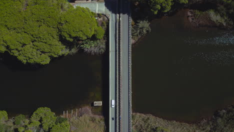 Modern-cars-driving-over-a-river-bridge-from-above-along-a-countryside-street-filmed-and-followed-in-bird-view-by-aerial-drone