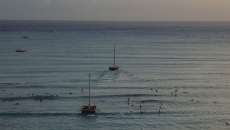 Two-Catamarans-Sailing-Towards-Sunset-Through-Group-of-Surfers-in-Waikiki-Bay,-Hawaii