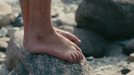 Man-standing-barefoot-at-top-of-the-rock-close-to-beach,-close-up