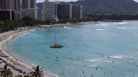 Yellow-Catamaran-Setting-Sail-against-Waves,-Surrounded-by-crowds-of-Beachgoers,-Hawaii,-Wide-Shot