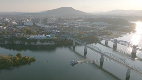 Weitläufige-Luftaufnahmen-Des-Tennessee-River-Mit-Der-Innenstadt-Von-Chattanooga-Und-Dem-Aussichtsberg-Im-Hintergrund-Und-Booten-Unter-Den-Brücken
