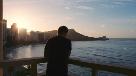 Silhouettierter-Tourist,-Der-Allein-Auf-Dem-Hotelbalkon-Steht-Und-Den-Wunderschönen-Morgensonnenaufgang-über-Der-Bucht-Von-Waikiki,-Hawaii,-Beobachtet