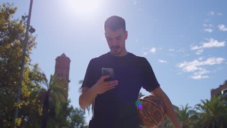 Nice-medium-shot-of-a-young-Caucasian-man-looking-at-his-cell-phone-while-playing-basketball-on-a-street-basketball-court-on-a-sunny-day-with-flares