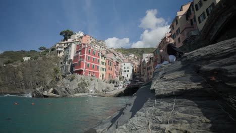 Female-Tourist-Sitting-On-Rock-By-The-Sea-With-Riomaggiore-In-Backdrop