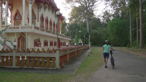 A-stationary-footage-of-a-lone-female-cyclist-walking-her-bike-beside-a-sacred-asian-temple