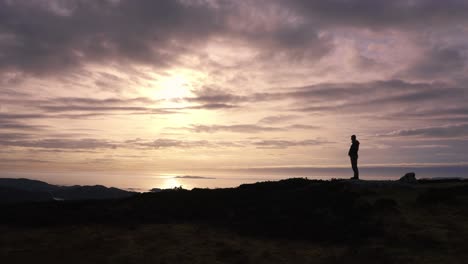 Man-silhouette-standing-in-front-of-striking-sunset-view-by-coastline-sea-on-Mull,-Scotland