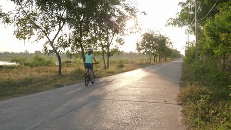 Una-Toma-Panorámica-Estacionaria-De-Una-Ciclista-Montando-Su-Bicicleta-A-Lo-Largo-De-Una-Calle-Vacía-En-Isan,-Tailandia