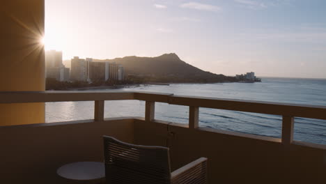 Slow-Motion-Pan-of-Waikiki-Bay-with-Sun-Flare-and-Empty-Chair-on-Hotel-Balcony,-Wide-Shot
