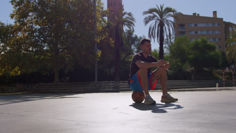 General-shot-of-a-young-Caucasian-man-sitting-on-top-of-a-basket-ball,-relaxed-eating-an-energy-bar-on-a-street-basketball-court-in-Barcelona,-Spain