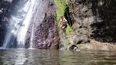 young-man-with-blond-hair-jumping-into-water-with-tropical-waterfall-behind-and-making-a-splash
