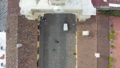Aerial-view-following-a-tuktuk-driving-under-the-Santa-Catalina-Arch-in-sunny-Antigua,-Guatemala---top-down,-drone-shot