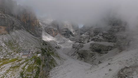 Fliegen-Durch-Graue-Wolken,-Die-Felsige-Berge-Und-Wanderhütten-In-Dolomiten-Enthüllen