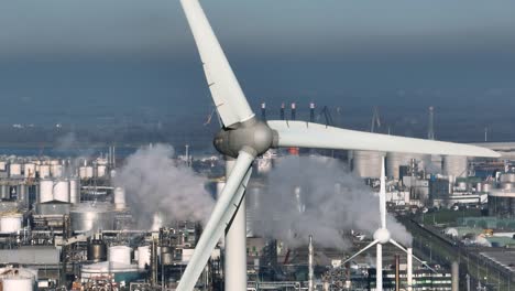 Close-up-aerial-orbiting-shot-of-a-wind-turbine-in-a-very-dense-industrial-zone-with-stacks,-steam-and-shiny-steel-tanks-in-the-background-on-a-sunny-day-near-Rotterdam-Netherlands