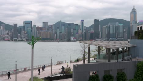 Leute-Werden-Gesehen,-Wie-Sie-Durch-Die-Uferpromenade-Von-Victoria-Harbour-Gehen,-Während-Sie-Ihren-Abend-Und-Die-Aussicht-Auf-Die-Skyline-Der-Wolkenkratzer-Der-Insel-Hongkong-Im-Hintergrund-Genießen