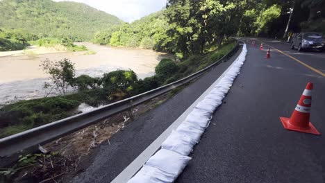 Sandsäcke-Und-Rote-Leitkegel-Säumen-Die-Straße-Entlang-Des-Fließenden-Flusses-Mit-Hohem-Wasserstand-Während-Der-Hochwassersaison-In-Thailand