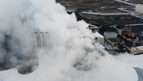 Thick-white-steam-rising-from-silica-pool-and-chimneys-at-geothermal-power-plant,-aerial