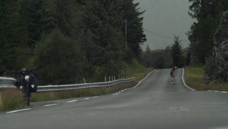 Skateboarder-taking-a-break-from-a-bike-trip-by-doing-tricks,-far-away-from-camera,-on-a-desolate-road-in-the-middle-of-nowhere-with-bad-weather-brewing-in-the-background