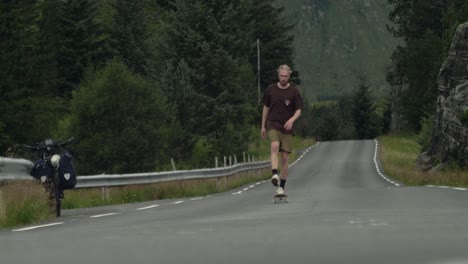 Young-caucasian-male-skateboarder-skateboarding-uphill-towards-camera-on-a-desolate-road-in-the-middle-of-nowhere-in-Norway,-scandinavia