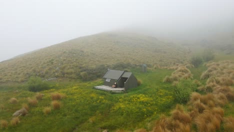 Hiker-relaxing-on-deck-of-a-hut-within-tussock-grass-covered-hills-in-New-Zealand