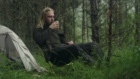 Young-long-haired,-bearded,-caucasian-man-solo-camping-in-the-woods,-sitting-drinking-coffe