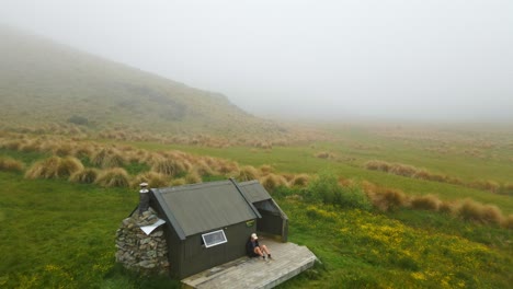 Aerial-dolly-revealing-hiker-sitting-in-front-of-secluded-mountain-hut-in-foggy-conditions-in-Canterbury,-New-Zealand