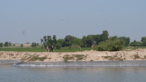 Schuss-Einer-Sprinkleranlage-Mit-Zentralem-Drehpunkt-Auf-Der-Anderen-Seite-Eines-Flusses-über-Ackerland-Mit-Einheimischen,-Die-In-Punjab-Vorbeiziehen