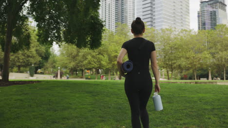 White-woman-in-black-clothing-walking-through-a-Seattle-park's-grass-with-her-yoga-mat