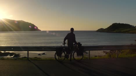 Young-male-arriving-at-a-beautifull-veiwpoint-with-fully-packed-bike-in-sunset
