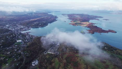 Aerial-descending-shot-through-hazy-clouds-mist-revealing-the-stunning-fishing-village-of-Oban,-Scotland,-Mull-Island-and-West-highlands-drone