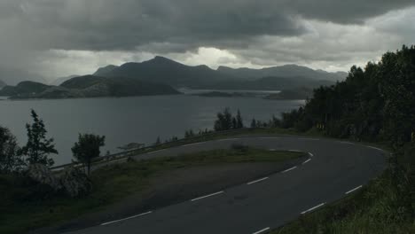 Young-man-cycling-uphill-in-dark,-moody,-windy-weather,-storm-brewing,-with-fully-packed-touring-bike