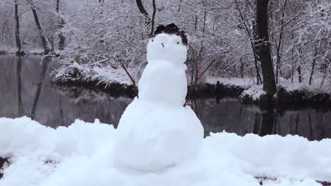 A-snowman-on-the-river-bank-in-the-middle-of-the-European-forest-with-gentle-snowfall---trucking-shot