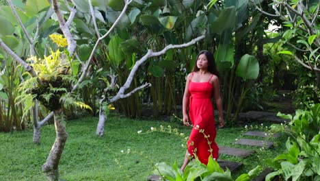 slow-motion-of-young-asian-female-walking-through-lush-green-garden-in-a-red-maxi-dress-on-sunny-day