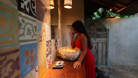 young-tan-asian-woman-putting-on-lipstick-and-makeup-in-traditional-outdoor-balinese-bathroom-with-mosaic-tile