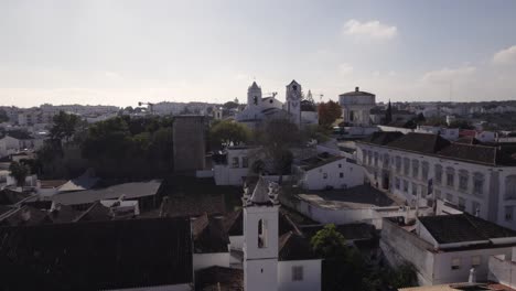 Aerial-orbiting-over-Tavira-cityscape,-with-the-Church-of-Santa-Maria-in-the-distance,-Sunny-day