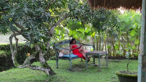 young-tan-woman-in-red-maxi-dress-sitting-on-a-bench-in-a-tropical-lush-garden-reading-a-book