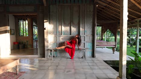 Junge-Asiatische-Frau,-Die-Auf-Holzbank-In-Bali-villa-Im-Joglo-stil-Mit-Einem-Schönen-Roten-Maxikleid-Posiert