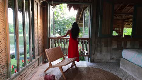Asiatische-Frau-Mit-Langem-Rotem-Kleid,-Die-Aus-Dem-Balkonfenster-In-Einer-Traditionellen-Böhmischen-Villa-Auf-Bali-Schaut