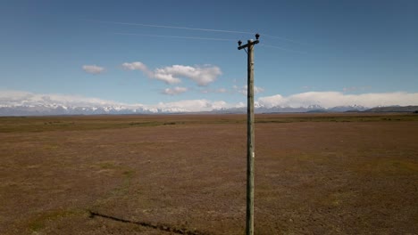 Aerial-footage,-passing-wooden-power-pole-closely-in-a-vast,-dry-landscape-with-mountains-in-background