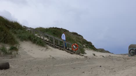 Handsome-White-male-walks-down-sand-dune-on-old-wooden-stairs-to-sandy-beach