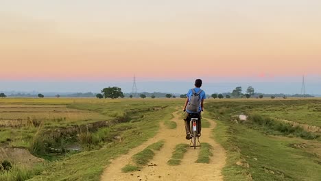 Young-village-boy-with-backpack-cycle-in-rural-path-through-agriculture-fields-in-Bangladesh,-back-view