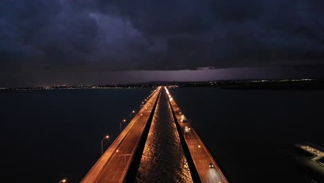Stationary-drone-shot-of-Bridge-and-Ocean-with-Storm-in-the-Distance,-lightning-striking-the-town-in-distance,-taken-at-night