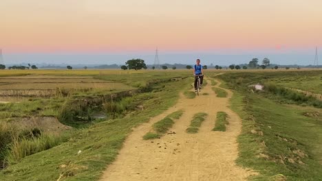Niño-De-Aldea-En-Bicicleta-Por-Un-Camino-De-Tierra-En-Las-Tierras-De-Cultivo-De-Bangladesh,-Vista-Frontal