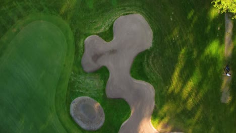 Drone-shot-over-golf-course-showing-bunkers,-trees,-and-players-during-a-sunny-day-and-golf-practice