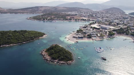 Orbiting-aerial-view-of-serene-waters,-boats,-lush-islands,-and-towering-mountain-views-by-the-charming-Ksamil-village-on-the-Ionian-Sea-coast